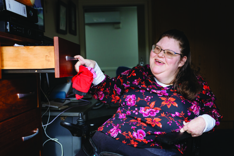 A person who uses a wheelchair is smiling and putting away their laundry in their bedroom.