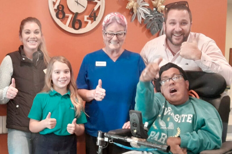 A group of people smiling and giving a thumbs up signal with their hands. One person uses a wheelchair.