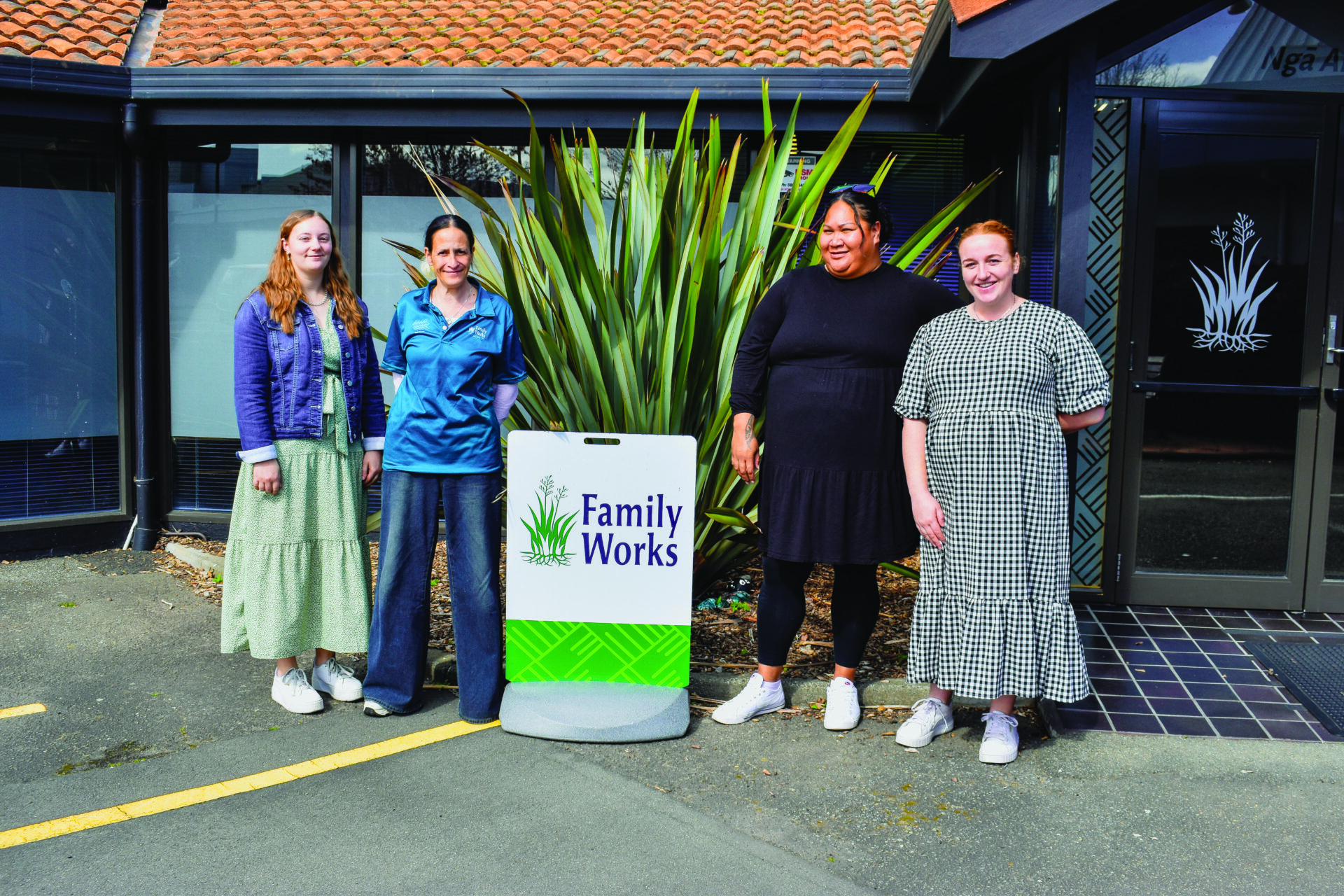 Some of the dedicated Family Works team supporting through our local schools. Four team members are standing outside the Family Works office in Hastings with a sign that says Family Works. There is a harakeke plant behind them.