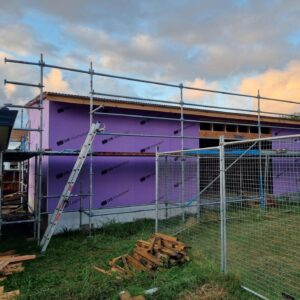 Outside view of a building in progress. Scaffolding surrounds the building and the roof is on. There are building materials in the foreground.