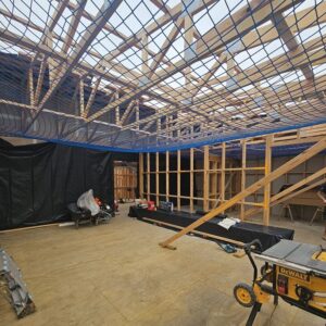 Inside view of a building in progress. Wooden floor and framing. The sky can be seen through the roof trusses. There is construction equipment in the foreground.