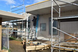 A painter is on a ladder painting the outside of the new Rowan Apartments. There is scaffolding around the building and the start of a wooden accesibility ramp. 