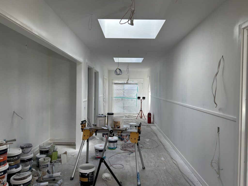 Inside a construction site showing neatly finished walls. Lots of paint tins are on the floor. There are cables hanging from walls and ceiling and a builder's light at the end of a passage.