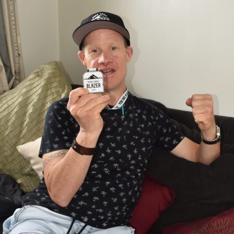 Peter Cullwick holds up his medal after completing the Peak Trail Blazer for charity. He is smiling and holding up a fist celebrating his great achievement.