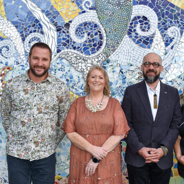 Three people stand smiling against a mosaic covered wall.