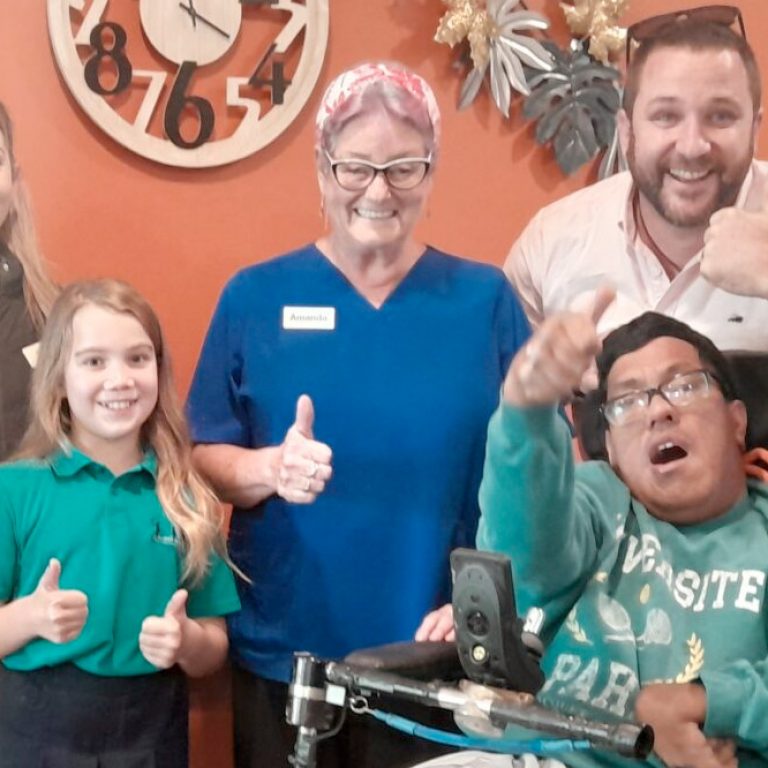 A group of smiling people including a nurse, a wheelchair user, a child and parent, and Enliven Disability Service Manager, Andrew Wordsworth. The group are giving a thumbs up signal with their hands.